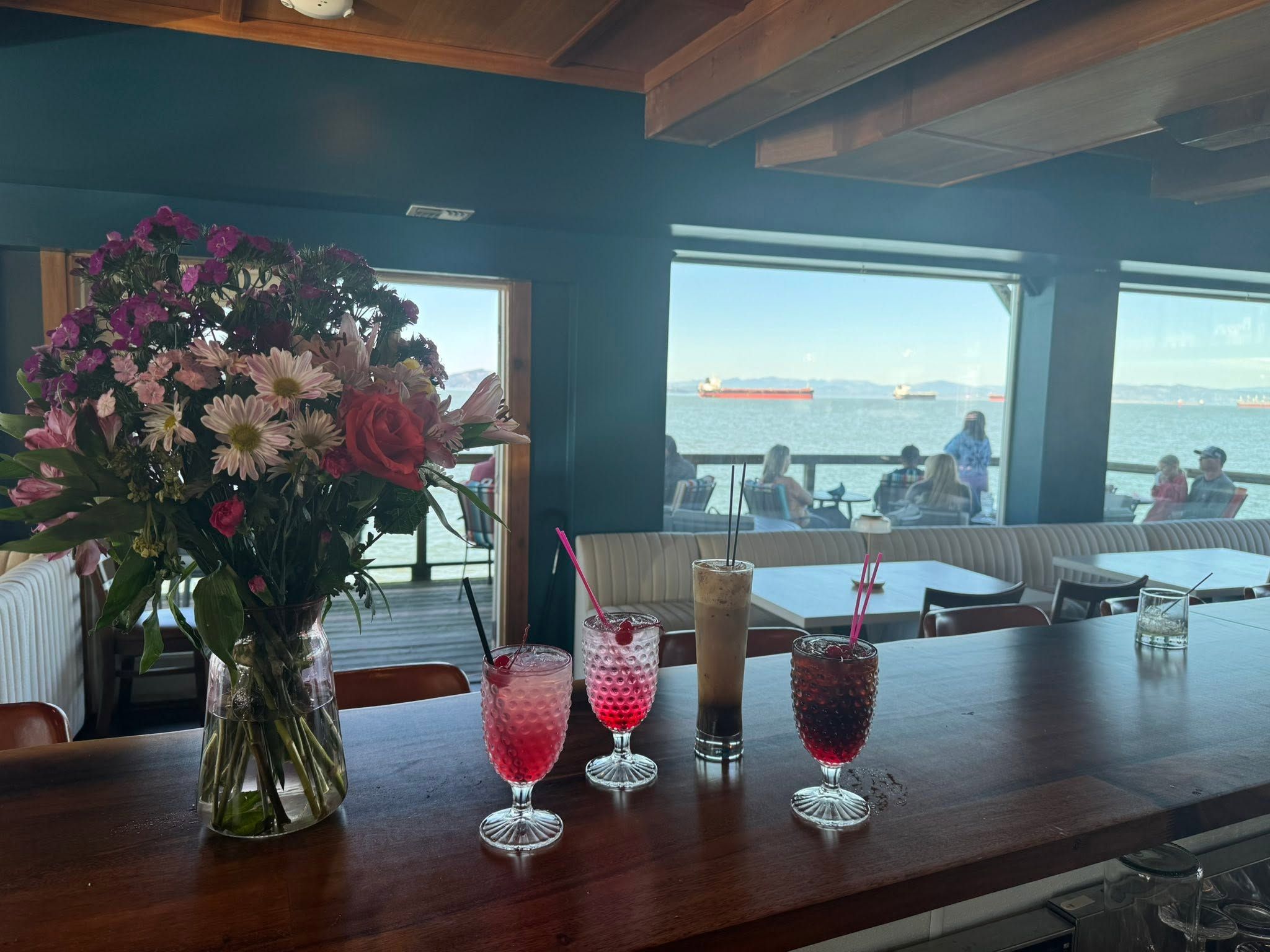 Looking out from inside Akres on the Pier: drinks and flowers on the counter, the deck beyond the windows, and ships at anchor on the Columbia River
