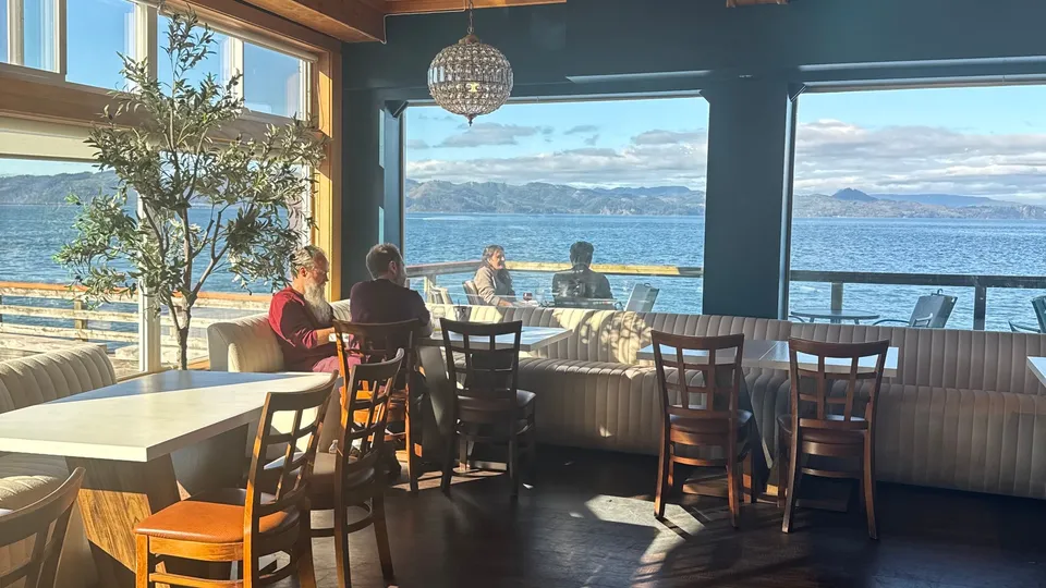 The main dining room in daylight: banquettes and wooden tables under a timber ceiling, with the river and the hills through the big windows