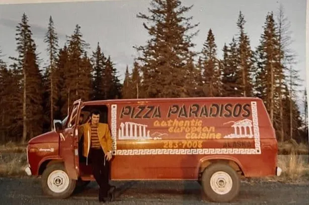 Konstantinos Macheras leaning on a red delivery van lettered Pizza Paradisos, authentic European cuisine, Alaska, with spruce trees behind