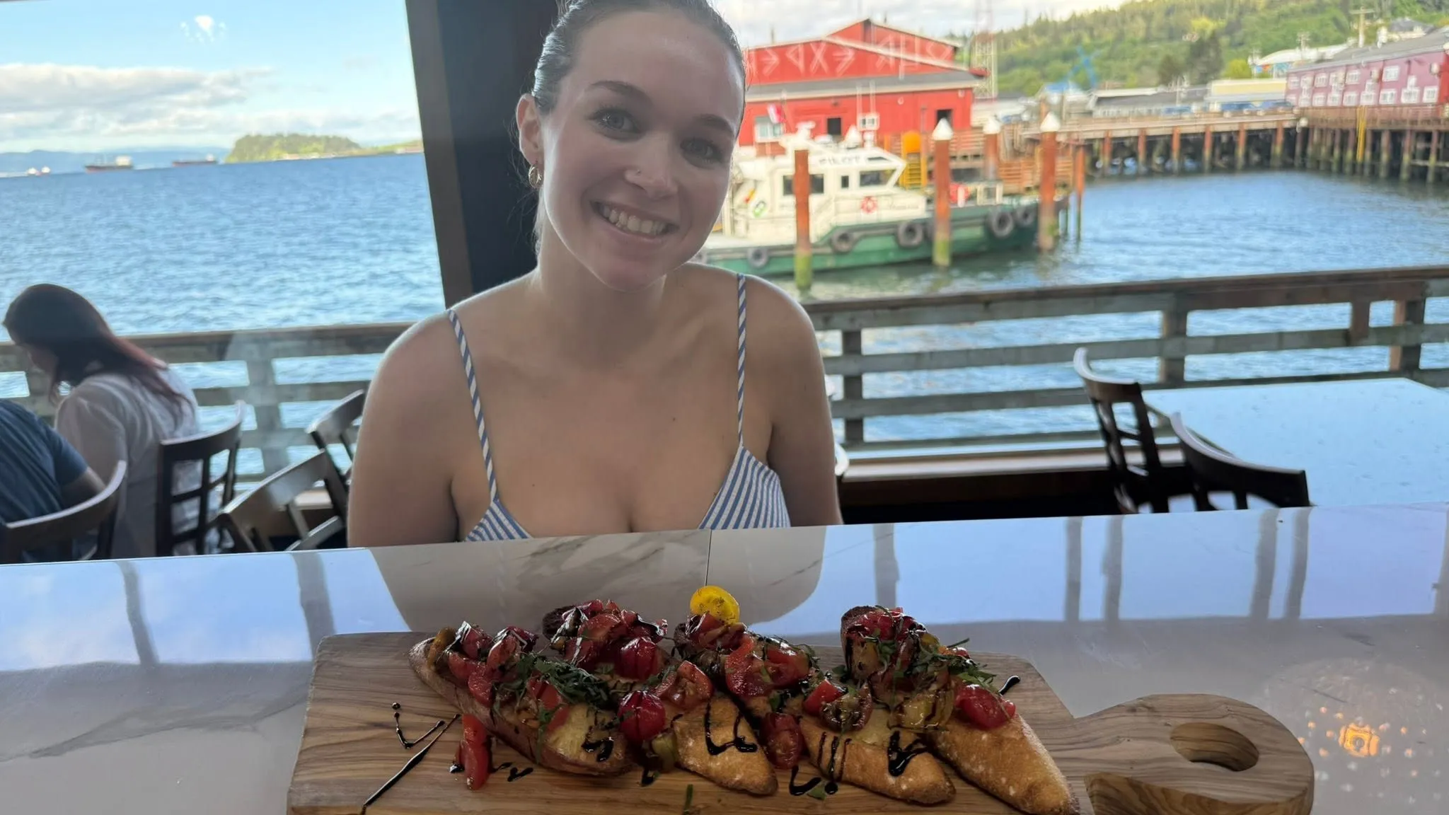 Samantha Macheras smiling at the kitchen's service window, a board of food ready to go out in front of her and the Astoria waterfront through the windows behind